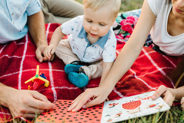 Happy family holiday on a summer day. Mother, father, and baby son sitting on a blanket on picnic in mountains.  Mom, dad play and hug child at sunset. Spend time together. Parents read books for kid.