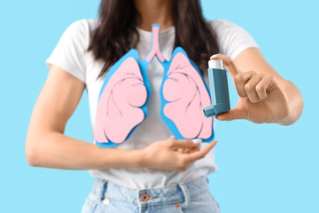 Young woman with paper lungs and asthma inhaler on blue background, closeup