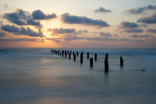 Wooden stumps from the base of an old, abandoned pier stretch out across the sea at sunset.