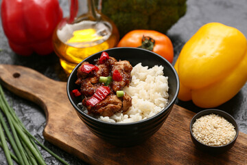 Tasty sweet and sour chicken with rice in bowl, vegetables and board on black grunge background, closeup