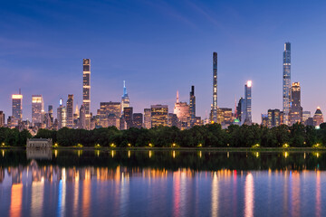 Naklejka premium New York City skyline at dusk with reflections of Billionaires Row supertall skyscrapers in Central Park Reservoir. Evening view of illuminated luxury towers in Midtown Manhattan