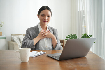 An Asian woman expresses gratitude during a virtual meeting from home, gently placing her hands on her chest while smiling at her laptop. She sits at a wooden desk in a bright, peaceful living room.