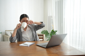 An Asian woman rubs her eyes while taking a coffee break during work from home. She sits at a wooden desk in a bright living room, appearing tired yet determined to stay productive.
