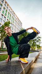 Young man, breakdancer in green shirt and black jacket executes stylish pose on ledge, showcasing...