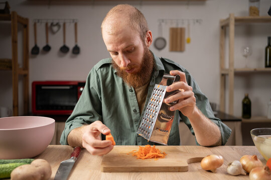 man grating fresh ripe carrot at wooden table in his home kitchen