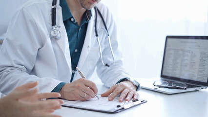 Doctor and a patient. The physician, wearing a white medical coat over a green shirt, is filling out a medical record form during a consultation in the clinic. Medical service