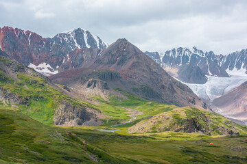 Vivid dramatic alpine scenery with green hills and rocks with view to high rocky pointy peak, sharp rockies, large snow-capped mountain top, snowy range and big glacier tongue under gray cloudy sky.