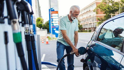 Cheerful mature businessman filling up car tank with fuel at gas station. Happy mid adult man...