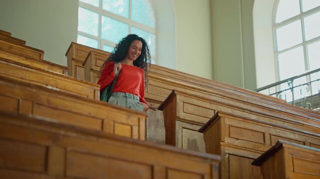 Smiling girl student Hispanic female Latina woman lady with backpack walk come in classroom university college big empty auditorium go down stairs after lecture class lesson learning study education