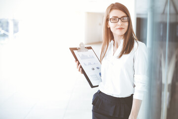 Beautiful female specialist with clipboard standing in sunny office and smiling charmingly. Business people concept