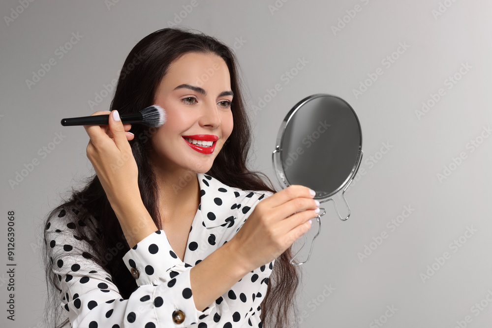 Wall mural smiling woman with mirror applying makeup on light grey background