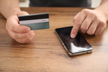 Man with credit card using smartphone at wooden table indoors, closeup
