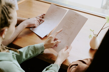Group of diners examining menu choices in cozy restaurant beside bright window, focusing on different options and discussing their preferences