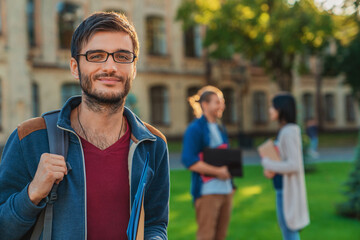 Caucasian male college student man guy in eyeglasses casual clothes with backpack holding books notebooks standing outside on modern university campus with friends classmates on background.