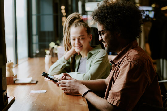 Smiling people relaxing in cozy cafe and using smartphones while enjoying coffee. Modern setting with natural light and friendly atmosphere enhancing experience