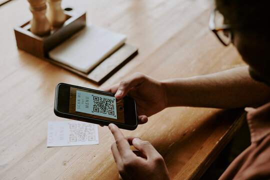 Close-up of hands scanning QR code with smartphone on wooden table, device visible in background displaying QR code, reflection of glasses seen in user's hand - Powered by Adobe