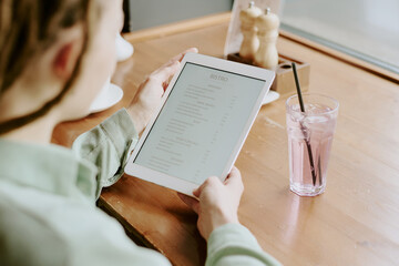 Person holding tablet reviewing digital menu in cozy bistro while drinking refreshment. Hands gripping device revealing tranquil dining setting with glass of beverage