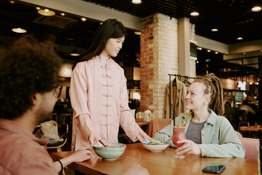 Asian waitress serving bowls of food to diverse group of customers in busy restaurant setting, engaging in friendly conversation, and creating welcoming atmosphere