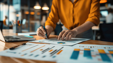 Fototapeta premium A person in a bright shirt is analyzing graphs and charts on a desk in an office environment. A laptop is open nearby with blurred background elements.