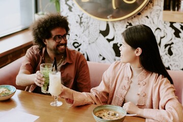 Biracial couple enjoying delicious Asian cuisine while having drinks at restaurant, smiling and talking joyfully. Both are seated at wooden table, natural light from window illuminating scene