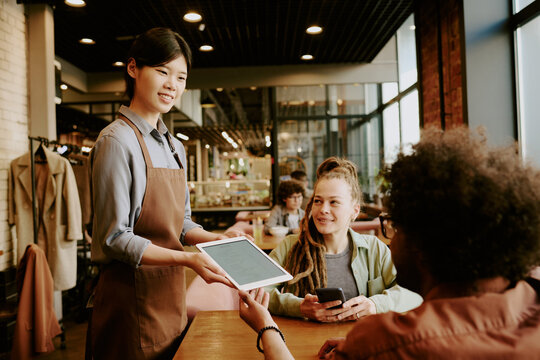 Chef holding digital menu beaming at diverse group of young customers in modern, stylish cafe with brick walls and large windows creating cozy atmosphere