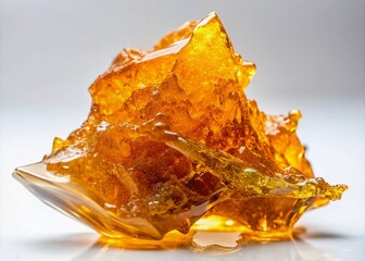 Golden amber shatter, a translucent cannabis concentrate, rests on a white background, its fragile glass-like texture and crystalline structure glistening with potency and purity.