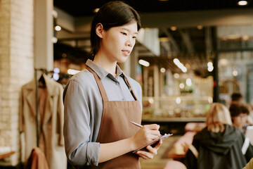 Young Asian woman wearing an apron taking notes in a busy restaurant surrounded by customers and staff working in background