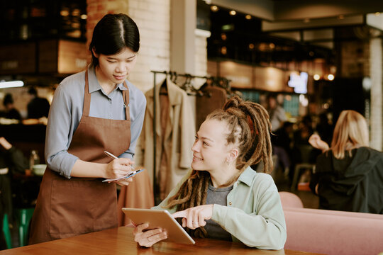 Asian waitress in apron taking order from smiling woman with tablet in warm cafe setting, engaging in friendly interaction and creating inviting atmosphere