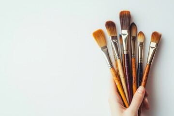 Minimalist photograph of a hand holding a selection of artist paintbrushes against a white background with shadows.