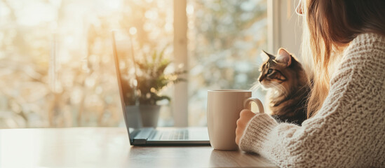 Obraz na płótnie Canvas A girl is sitting at a table by the window, holding a mug of tea and looking at a laptop monitor, a cat is sitting next to her and also looking at the monitor. An empty space for the text.