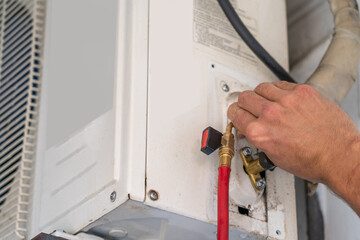 Technician's hand adjusting connections on outdoor air conditioner unit with red hose attached for maintenance and freon refill. Professional HVAC system repair and service, ensuring optimal cooling