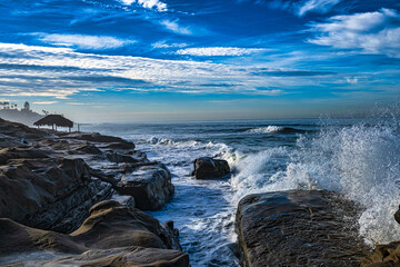 2023-12-31 ROCKY SHORELINE AT THE WINDANSEA BEACH WITH A GRASS HUT AND WAVES CRASHING ON THE ROCKS SPRAYING WATER IN LA JOLLA CALIFORNIA NEAR SAN DIEGO