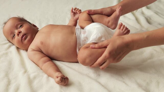 Anonymous mother doing gymnastics with her newborn child to relieve gases and alleviate constipation moving kid's legs infant lying on bed on white sheet