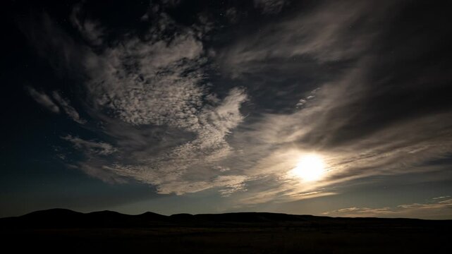Time lapse of thin clouds moving through the night sky in front of a full moon.  Scattered stars can be seen in the darker portion of the sky.  Small rolling hills define the horizon.
