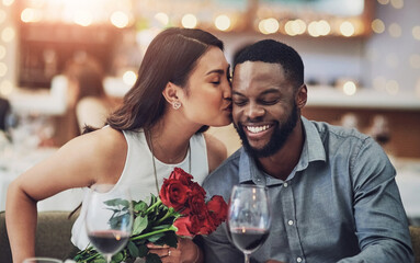 Couple, dinner and kiss on date in restaurant for flowers, romance or commitment to relationship. Interracial, people and happy with celebration for anniversary, honeymoon or valentines day with wine