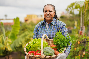 Happy african senior woman smiling on camera while holding basket with fresh vegetables from house...