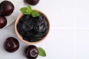 Delicious prunes with mint in bowl and fresh ripe plums on white tiled table, flat lay. Space for text