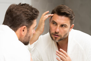 a man in a bathrobe in the bathroom looking at his wrinkles and pimples on his face. A man is looking at his reflection in a mirror. A man is looking at his reflection in a mirror. 