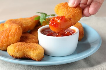 Woman dipping tasty chicken nugget into chili sauce at grey table, closeup
