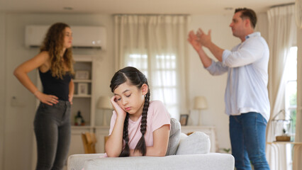 Annoyed and unhappy young girl sitting on sofa trapped in middle of tension by her parent argument in living room. Unhealthy domestic lifestyle and traumatic childhood develop to depression Synchronos