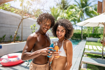 Young couple of African descent enjoying a sunny day at a luxurious poolside, laughing and toasting with colorful drinks. They are dressed in swimwear, surrounded by a tropical and relaxing atmosphere