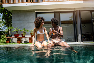 A young couple of African descent enjoying a sunny day at a luxurious poolside, sitting at the...