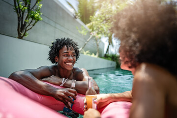 A young couple enjoying a relaxing moment in a pool, floating on a pink inflatable ring, and holding drinks. The backdrop features a modern villa and lush tropical palm trees. A perfect vacation.