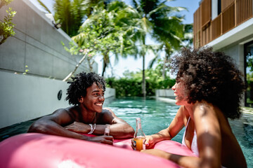 A young couple enjoying a relaxing moment in a pool, floating on a pink inflatable ring, and holding drinks. The backdrop features a modern villa and lush tropical palm trees. A perfect vacation.