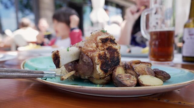 Close-up of roasted pork knuckle with crispy skin and roasted potatoes on a plate at an outdoor restaurant, with blurred diners in the background