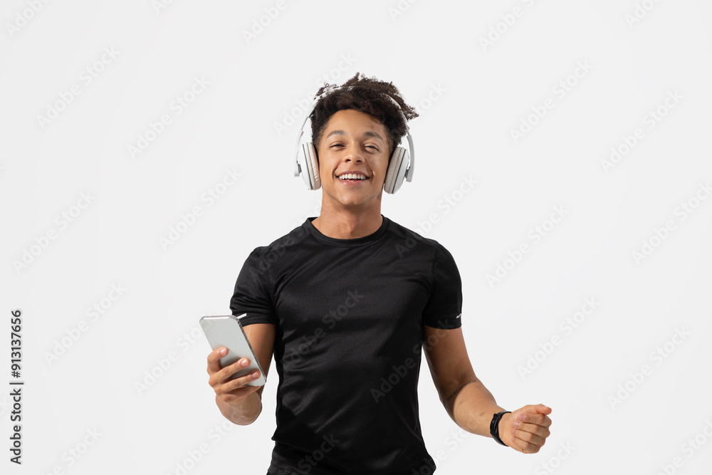 Wall mural A young man wearing headphones smiles joyfully while holding a smartphone in his hand. He is dancing in a relaxed manner, showcasing his enjoyment of music in a bright and simple environment.