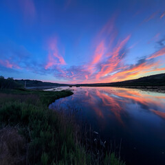  A vibrant sunset casts colorful reflections on a river, surrounded by lush green grass.