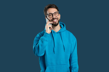 A young man dressed in a blue hoodie smiles bright while speaking on his phone. The warm expression on his face suggests he is engaged in a pleasant conversation