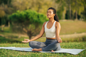 A woman sits cross-legged on a yoga mat in a tranquil park, practicing meditation. Surrounded by lush greenery and softly illuminated by sunlight, she radiates peace and focus.