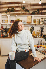 A cute woman in a white knitted sweater is sitting with coffee in a cozy kitchen with Christmas decorations. Kitchen in New Year's style.